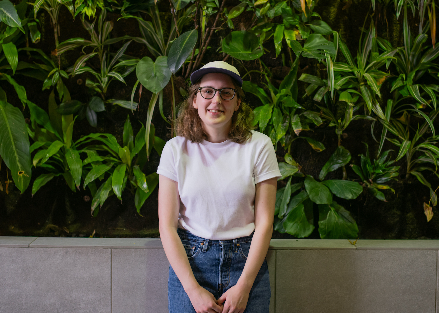 Laura stands in front of a living wall wearing a white shirt, blue jeans, glasses and a white baseball hat